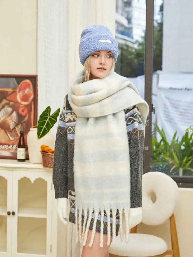 Woman wearing a grey knit sweater with blue and white geometric pattern, a light blue beanie, and a chunky striped scarf, looking to the side.