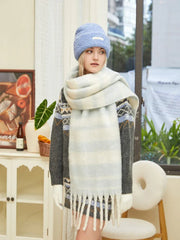 Woman wearing a grey knit sweater with blue and white geometric pattern, a light blue beanie, and a chunky striped scarf, looking to the side.