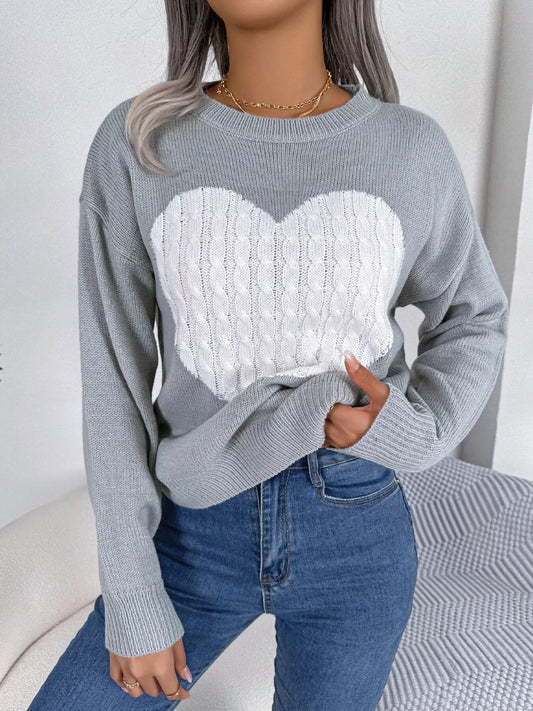 Woman wearing a grey knit sweater with a white cable knit heart graphic on the front, paired with blue jeans and gold necklaces.