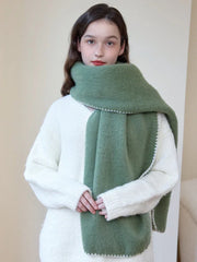 Woman wearing an oversized green scarf with a white fringed edge and a cream-colored sweater, with hands clasped in front.