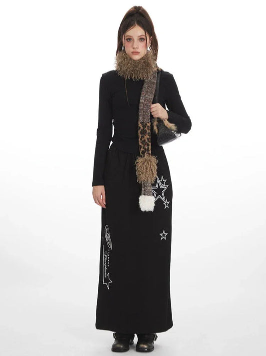 Model wearing a black long-sleeve top and a black maxi skirt with a drawstring waist and white star graphics, accessorized with a multi-patterned scarf and a black handbag.
