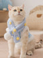A cream-colored cat wearing a light blue striped vest with white faux fur trim and yellow embroidered stars, accessorized with a matching light blue scarf with yellow stars and white pom-poms.