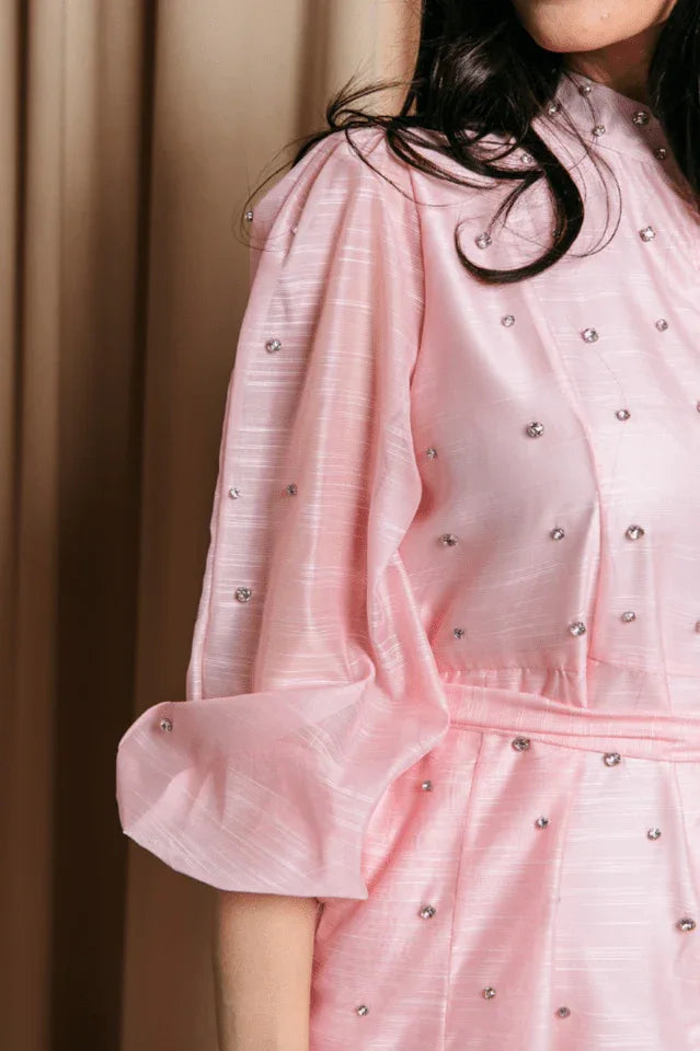 Close-up detail of the sleeve and bodice of a pink midi dress. The fabric has a subtle striped texture and is adorned with small, sparkling embellishments. The sleeve is a puff sleeve style.