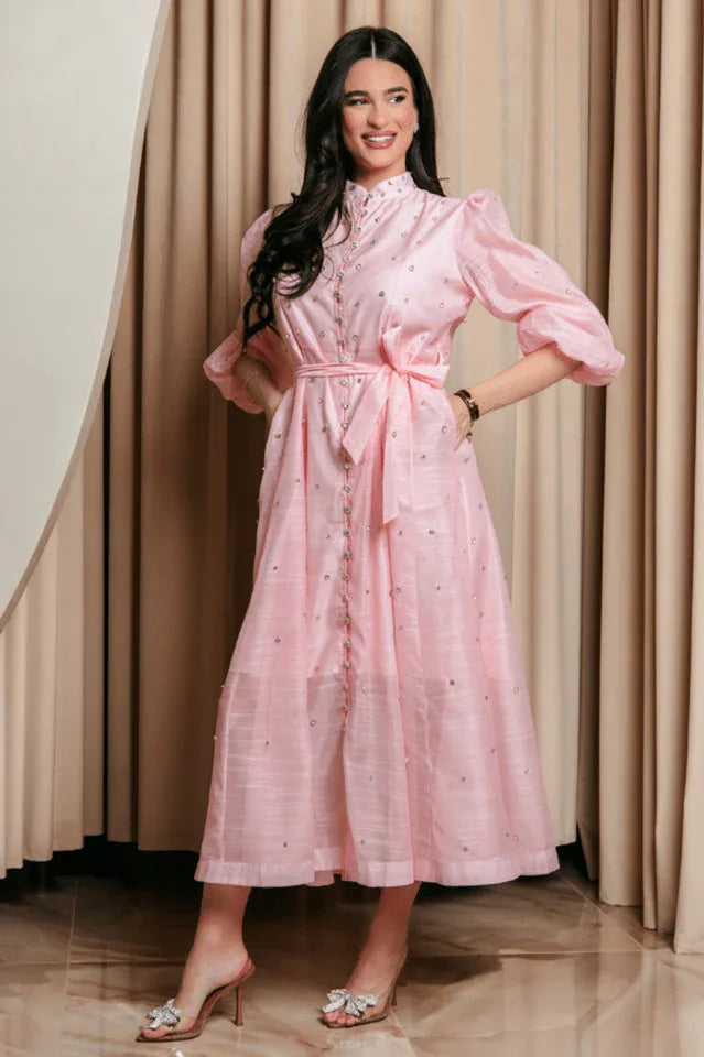 Full-length shot of a woman modeling a pink, embellished midi dress with puff sleeves and a belted waist. She has her hands on her hips and is wearing clear, bow-embellished heels.