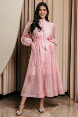 Woman wearing a pink, long-sleeved, midi dress with a high neckline, belted waist, and sparkling embellishments. She is standing with her hands in her pockets and wearing clear, bow-embellished heels.