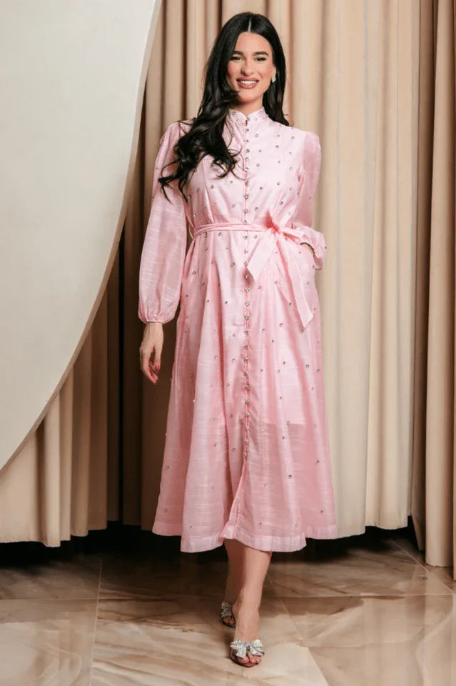 A woman stands in a pink, button-down midi dress adorned with scattered embellishments. The dress features long puff sleeves, a high neckline, and a matching belt. She is wearing open-toed heels with bow details.