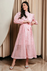 A woman poses in a pink, embellished midi dress. The dress has a high neckline, button-down front, belted waist, and long puff sleeves. She is wearing bow-embellished heels.
