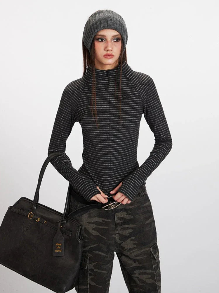 Close-up front view of a model wearing a gray striped long-sleeve top with a mock neck and thumbholes, paired with camouflage cargo pants and a black belt, holding a black tote bag.