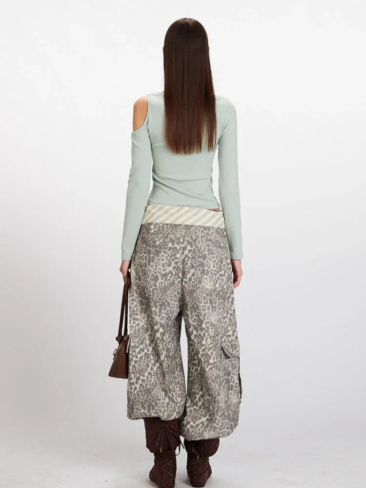 Back view of a woman wearing a mint green long-sleeved shirt with an asymmetrical neckline and a cut-out shoulder, paired with wide-leg leopard print pants, a striped belt, and brown boots.
