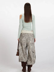 Back view of a woman wearing a mint green long-sleeved shirt with an asymmetrical neckline and a cut-out shoulder, paired with wide-leg leopard print pants, a striped belt, and brown boots.