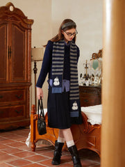 Full body shot of a woman in a layered outfit including a sweater, collared shirt, and pleated skirt, accessorized with a striped scarf and boots.