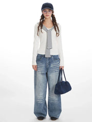 Model wearing a white long-sleeved top with striped trim and wide-leg blue jeans, holding a navy handbag and wearing a navy baseball cap.