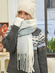 Woman adjusting her multi-colored fringed scarf while wearing a cream beanie and a gray knit sweater dress with Fair Isle pattern.