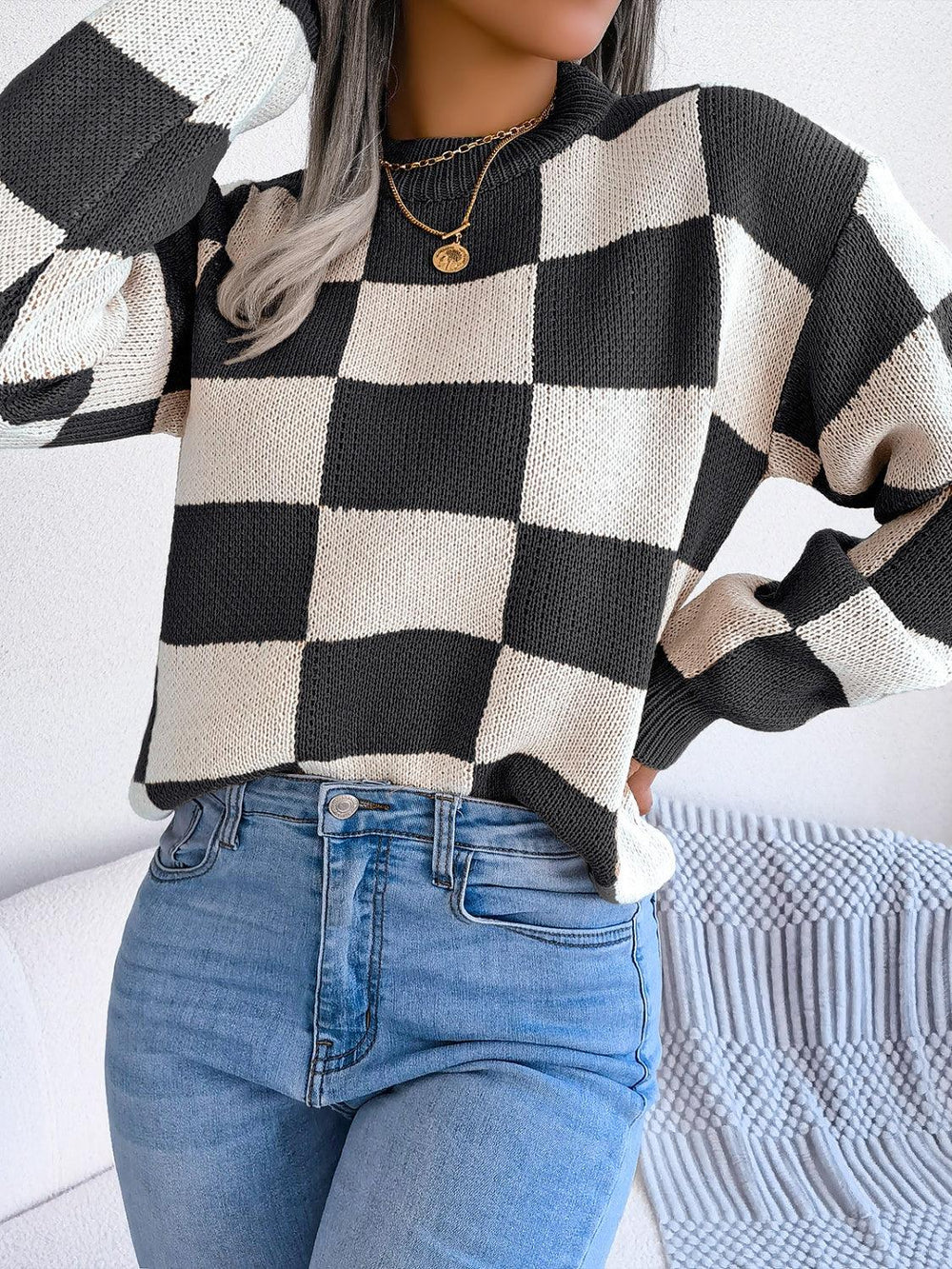 Woman wearing a black and white checkerboard knit sweater with light wash jeans, hands on hips, front view.