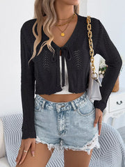 Woman wearing a black cropped knit cardigan with a tie front closure and denim shorts, holding a white handbag with a gold chain strap.