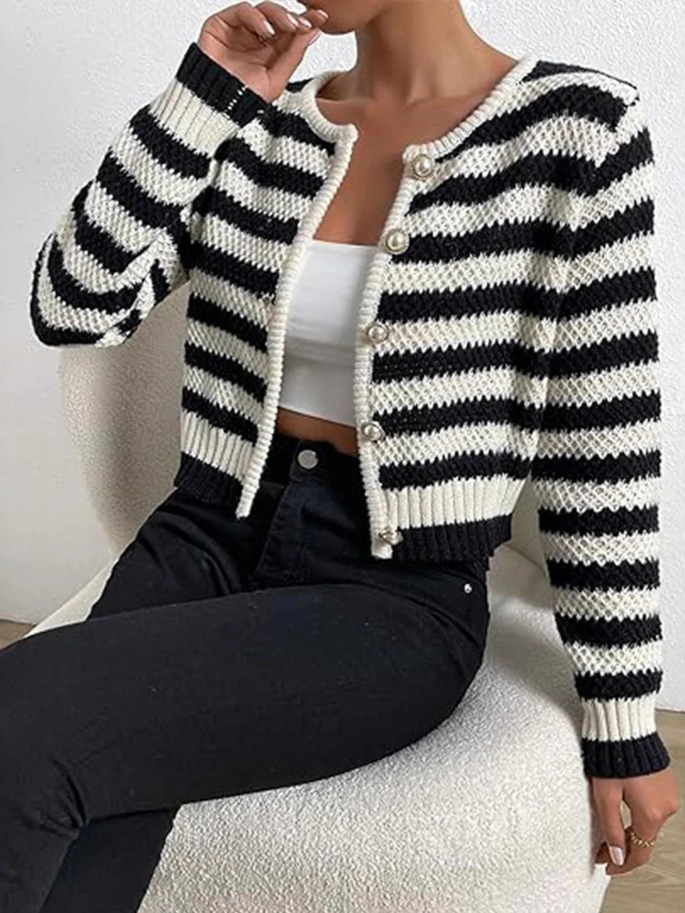 Woman wearing a black and white striped cropped knit cardigan closed over a white camisole and black jeans, seated on a chair.