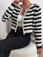 Woman wearing a black and white striped cropped knit cardigan closed over a white camisole and black jeans, seated on a chair.