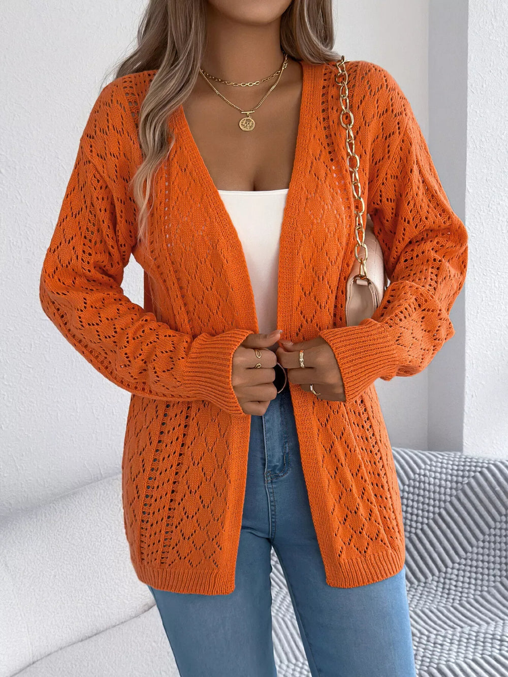 Woman adjusting an orange open-front knit cardigan, revealing a white tank top and blue jeans.