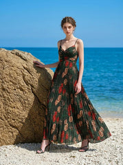 Woman wearing a dark green floral print maxi dress with spaghetti straps and a pleated skirt, standing on a beach with a large rock and the ocean.