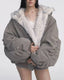 Model wearing a grey bomber jacket with white faux fur lining, hands tucked inside the jacket, white lace-trimmed camisole, and grey shorts.
