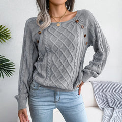 Woman wearing a grey cable knit sweater with buttons, paired with light wash jeans, standing in front of a white wall with a plant.