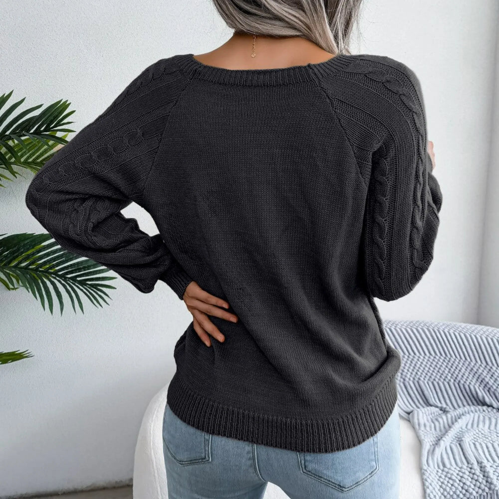 Back view of a woman wearing a charcoal grey cable knit sweater with detailed sleeves, hands on hips.