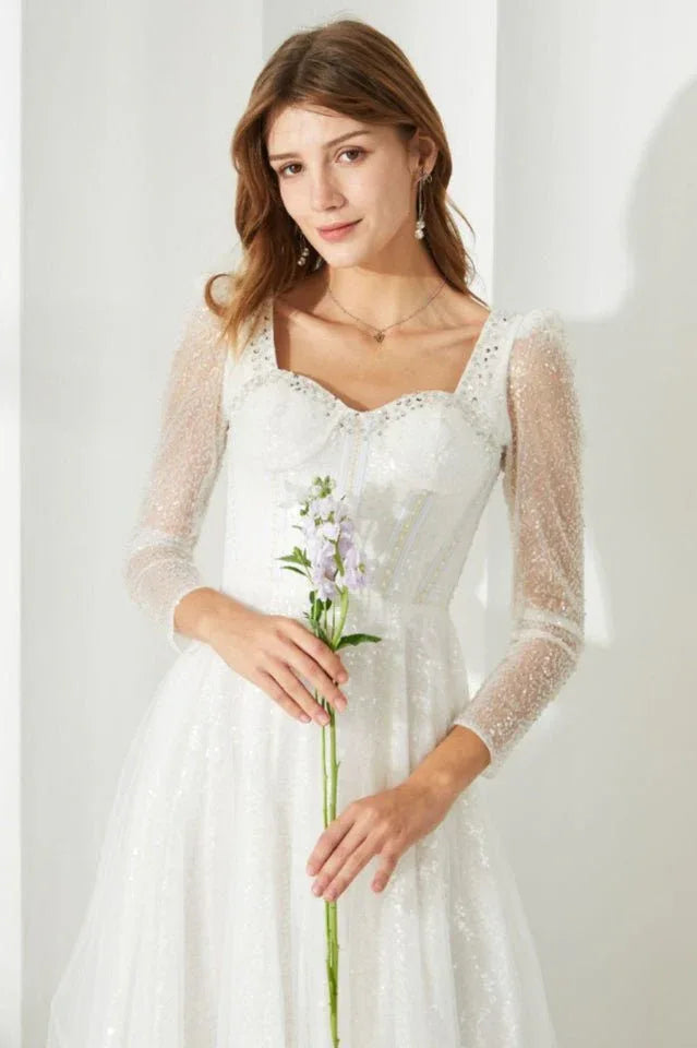 Close-up of a woman holding a flower, wearing a white sequined dress with a square neckline and sheer sleeves.