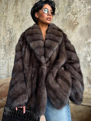 woman with short black hair wearing a brown fur coat with wide lapels and long sleeves, holding a black clutch