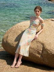 Woman wearing a floral print midi slip dress with off-the-shoulder sleeves and a cowl neckline, sitting on a rock by the sea.