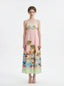 Full front view of a woman wearing a pink midi dress with a tropical print and spaghetti straps, standing against a white background.
