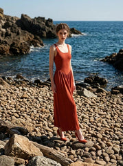 woman wearing a rust-colored ribbed knit midi dress with a scoop neckline and sleeveless design, posing on a rocky beach with the ocean in the background