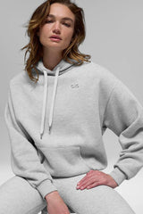 Close-up of a woman wearing a light grey cropped hoodie with a drawstring hood and embroidered logo.