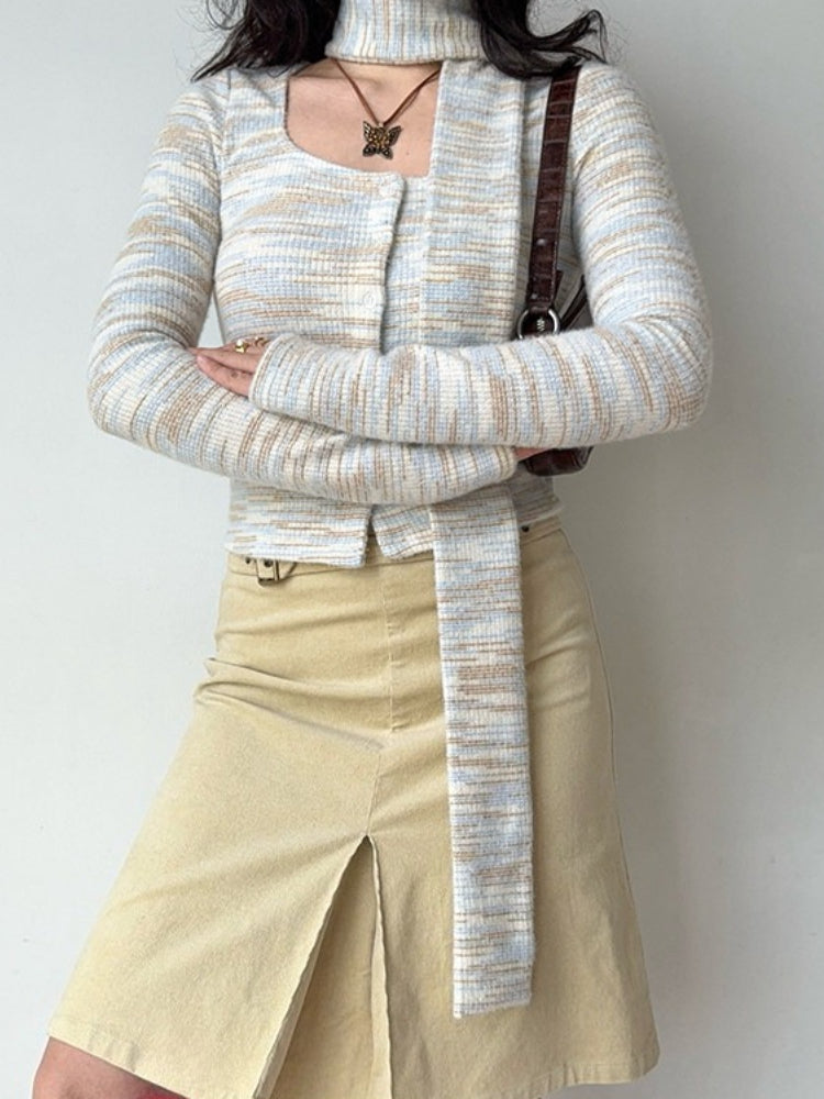 Woman with arms crossed wearing a light blue, white, and tan striped knit cardigan with a square neckline and an attached scarf, paired with a tan skirt.