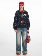 Full-length view of a woman wearing a navy blue hooded zip-up jacket with a white paw print pattern, a red t-shirt, light wash wide-leg jeans, a red baseball cap, and a red handbag.