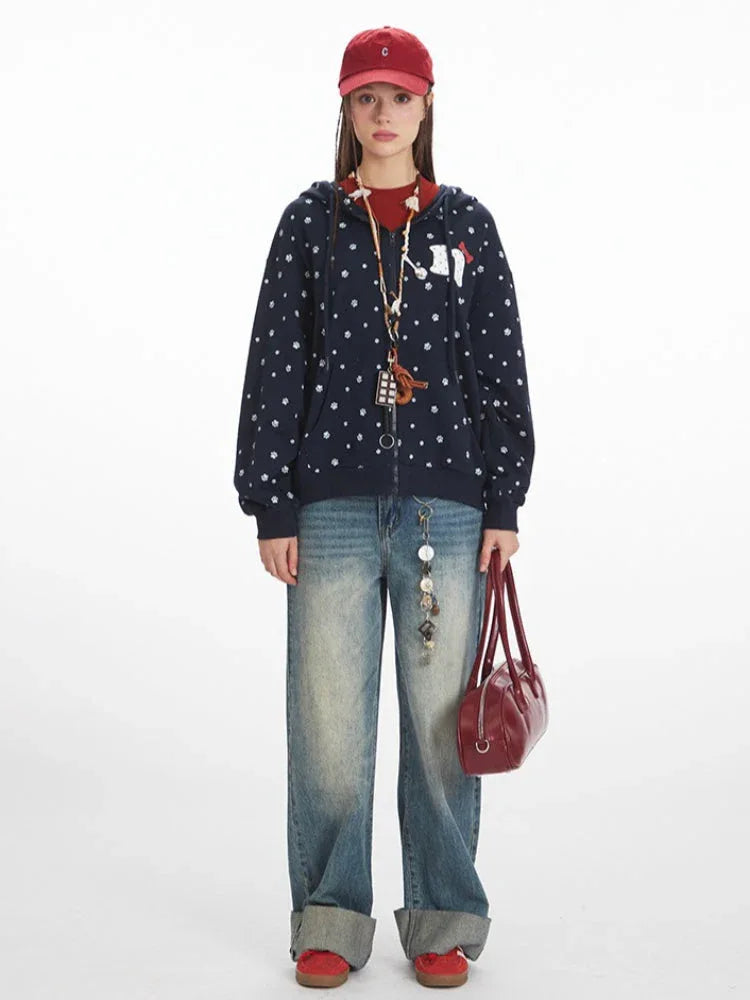 Full-length view of a woman wearing a navy blue hooded zip-up jacket with a white paw print pattern, a red t-shirt, light wash wide-leg jeans, a red baseball cap, and a red handbag.