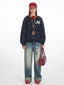 Full-length view of a woman wearing a navy blue hooded zip-up jacket with a white paw print pattern, a red t-shirt, light wash wide-leg jeans, a red baseball cap, and a red handbag.