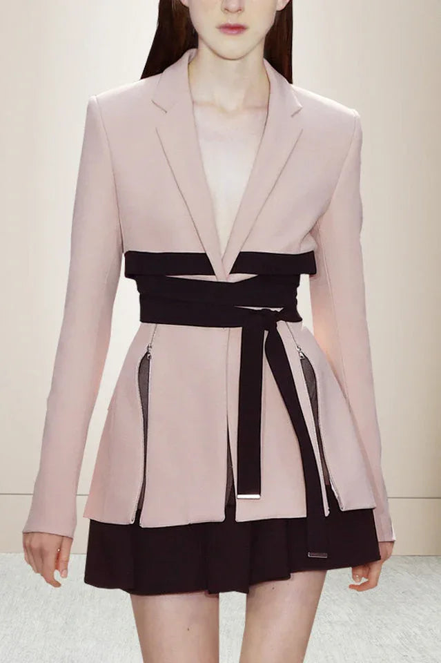 Close-up of a light pink blazer with a black wrap belt and black skirt, showcasing zipper details. Main image