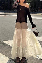 Model wearing a black off-the-shoulder top and a tiered maxi skirt with a brown pinstripe waistband, standing on a city street.