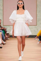 Model on a runway wearing a white organza mini dress with puff sleeves, a square neckline, and white sneakers.