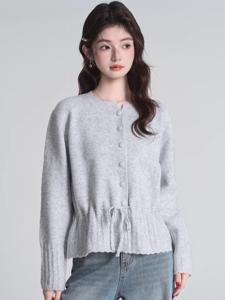 Front view of a woman modeling a light grey knit cardigan with a drawstring waist and button front closure, paired with blue jeans.