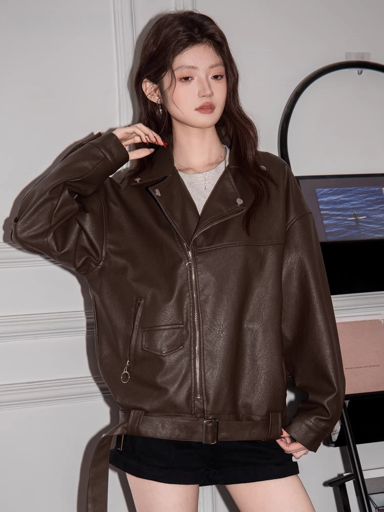 Woman wearing a brown faux leather moto jacket with a grey crop top and black shorts.