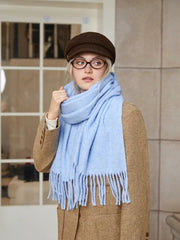 A young woman adjusts her light blue fringed scarf while wearing a brown tweed jacket.