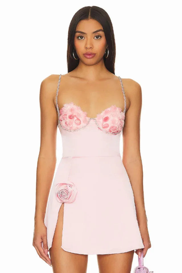 A woman models a pink mini dress with floral applique on the bodice and a high slit.