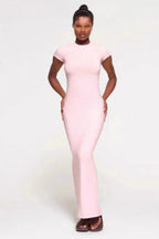 Woman wearing a light pink full-length ribbed knit dress with short sleeves and a crew neck, standing with hands on hips against a white background.