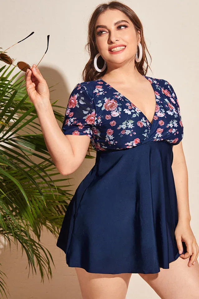 Plus-size woman in a navy blue floral print one-piece swimsuit with short sleeves and a skirted bottom, holding sunglasses.