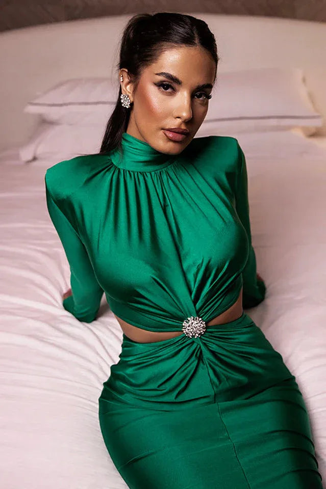Close-up of a woman wearing a long-sleeved green satin dress with a cut-out waist and embellished detail, sitting on a bed.
