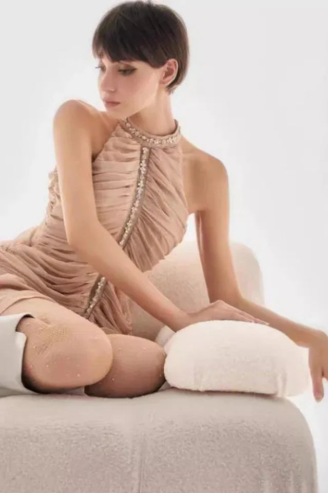 woman in a beige ruched halter neck mini dress with embellished neckline sitting on a chair Secondary image