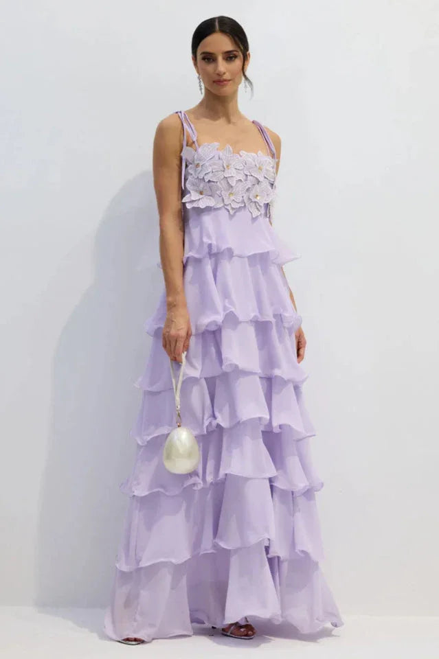 Woman wearing a long, light purple tiered ruffled chiffon dress with a floral applique bodice and tie shoulder straps, holding a pearl clutch. Main image