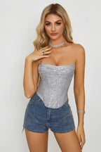 Woman in a silver sequined corset top and denim shorts, posing with hand on chest