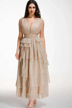 Woman wearing a floor-length, sleeveless, champagne-colored sparkly tiered gown with a V-neckline and empire waist.
